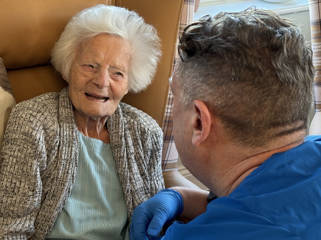 Photo shows a smiling care home resident sitting down in a high backed chair. A dentist is crouched down facing her as they chat.