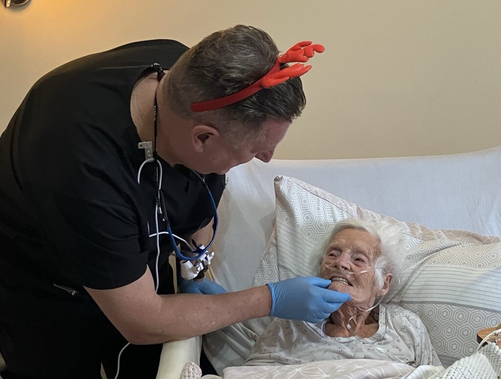 A dentist looks at the teeth of an elderly lady, as she reclines in a bed. 