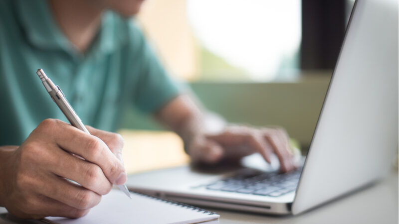 Man looking at a laptop holding a pen over a notepad